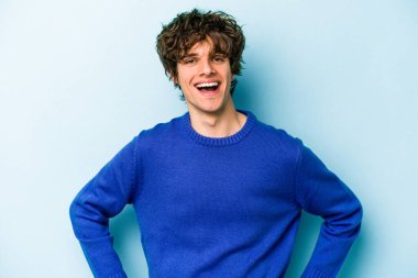 Kafkasyalı genç, mavi arka planda izole edilmiş. Kendine güvenen, kalçaları tutan biri..