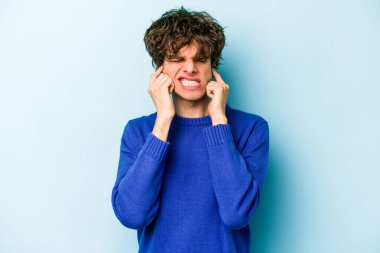 Mavi arka planda izole edilmiş genç beyaz adam kulaklarını elleriyle kapatıyor..