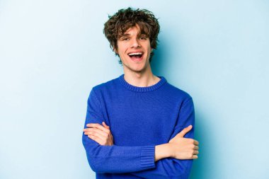 Mavi arka planda kendini güvende hisseden, kararlılıkla kollarını kavuşturan genç beyaz bir adam..