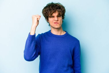 Mavi arka planda izole edilmiş genç beyaz adam yumruğunu kameraya gösteriyor, agresif yüz ifadesi var..