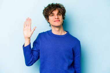 Mavi arka planda izole edilmiş genç beyaz adam gülümseyerek beş numarayı parmaklarıyla gösteriyor..