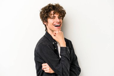 Beyaz arka planda izole olmuş genç beyaz bir adam gülümsüyor ve kendinden emin, çenesine elleriyle dokunuyor..