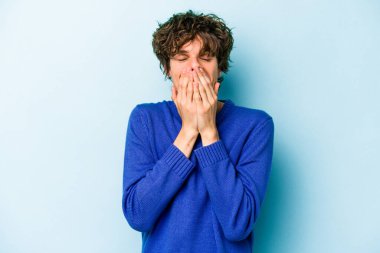 Mavi arka planda izole edilmiş genç beyaz bir adam bir şeye gülüyor, ağızlarını ellerle kapatıyor..