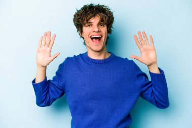 Mavi arka planda izole edilmiş genç beyaz adam on numarayı elleriyle gösteriyor..