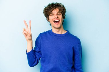 Kafkasyalı genç adam mavi arka planda izole edilmiş neşeli ve kaygısız parmaklarıyla barış sembolü gösteriyor..