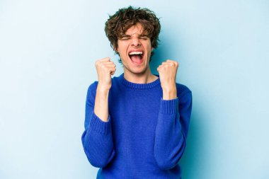 Kafkasyalı genç adam mavi arka planda izole edilmiş kaygısız ve heyecanlı tezahüratlar yapıyor. Zafer konsepti.
