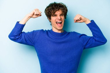 Mavi arka planda izole edilmiş genç beyaz adam güçlü bir hareket sergiliyor kollarıyla, kadınsı gücün sembolü.