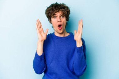 Mavi arka planda izole edilmiş genç beyaz bir adam zaferi ya da başarıyı kutluyor, şaşırıyor ve şok oluyor..