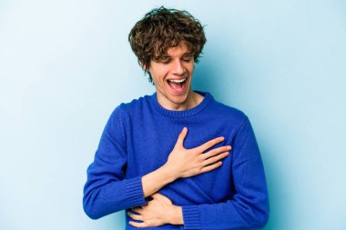 Mavi arka planda izole edilmiş genç beyaz adam mutlu bir şekilde gülüyor ve ellerini karnının üstünde tutarak eğleniyor..