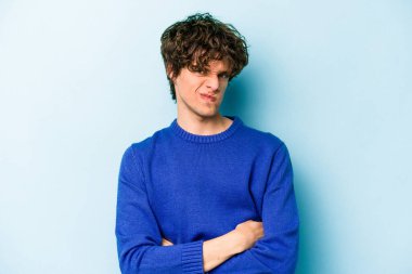 Mavi arka planda izole edilmiş genç beyaz adam alaycı bir ifadeyle kameraya bakarken mutsuz oluyor..