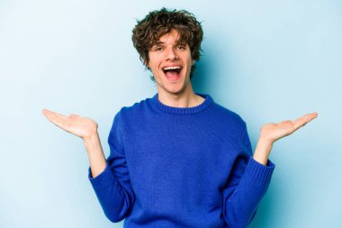 Mavi arka planda izole genç beyaz adam kollarıyla ölçek yapar, mutlu ve kendinden emin hisseder..