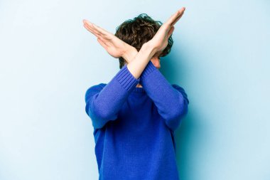 Kafkasyalı genç adam mavi arka planda izole edilmiş iki kolu çapraz tutuyor, inkar kavramı.