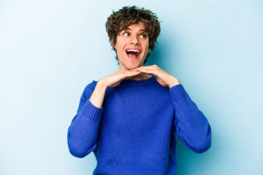 Mavi arka planda izole edilmiş genç beyaz adam ellerini çenesinin altında tutuyor, mutlu bir şekilde yana bakıyor..