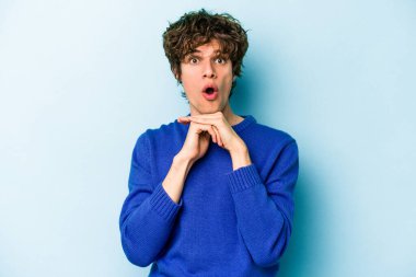 Kafkasyalı genç adam mavi arka planda izole edilmiş şans için dua ediyor, şaşırmış ve ağzını açıp önüne bakıyor..