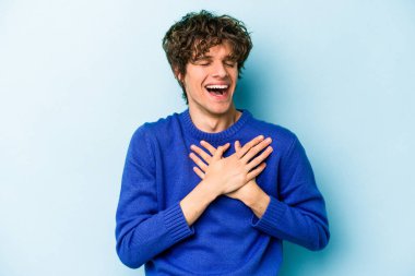 Kafkasyalı genç adam mavi arka planda izole olmuş gülüyor, elini yüreğine koyuyor, mutluluk kavramını koruyor..