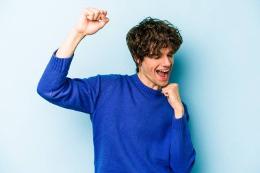 Mavi arka planda izole genç beyaz adam özel bir günü kutluyor, atlıyor ve enerjiyle kollarını kaldırıyor..