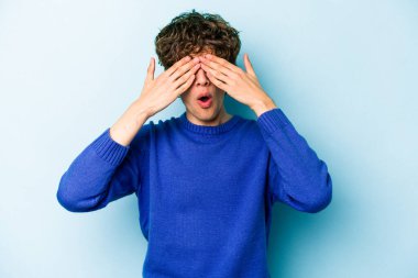 Mavi arka planda izole edilmiş genç beyaz adam elleriyle gözlerini kapatmaktan korkuyor..
