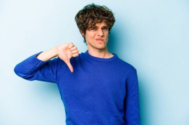 Mavi arka planda izole genç beyaz adam baş parmağını aşağı gösteriyor, hayal kırıklığı konsepti.