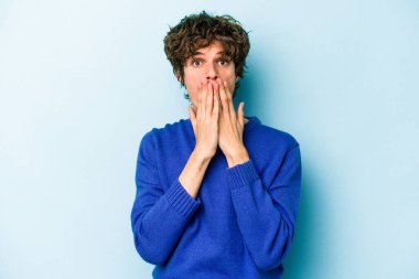 Mavi arka planda izole edilmiş genç beyaz adam şok olmuş, ağzı ellerle kaplanmış, yeni bir şey keşfetmeye hevesli..