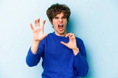 Mavi arka planda izole edilmiş genç beyaz adam pençelerini kedi taklidi yaparken gösteriyor, agresif bir hareket..