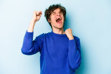 Mavi arka planda izole edilmiş genç beyaz bir adam zaferi, tutkuyu ve coşkuyu kutluyor, mutlu bir ifade..