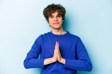 Kafkasyalı genç adam mavi arka planda izole edilmiş dua ediyor, bağlılık gösteriyor, dindar biri ilahi ilham arıyor..