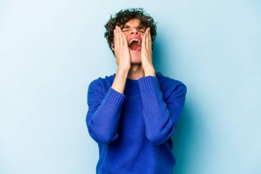 Kafkasyalı genç adam mavi arka planda izole edilmiş, ağlıyor, bir şeyden memnun değil, acı ve kafa karışıklığı konsepti..
