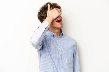 Beyaz arka planda soyutlanmış beyaz tenli genç bir adam elleriyle gözlerini kapatıyor, geniş bir gülümsemeyle bir sürpriz bekliyor..