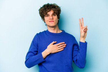 Kafkasyalı genç adam mavi arka planda izole edilmiş yemin ediyor, elini göğsüne koyuyor..