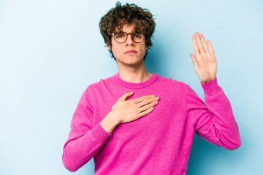 Kafkasyalı genç adam mavi arka planda izole edilmiş yemin ediyor, elini göğsüne koyuyor..