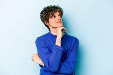 Kafkasyalı genç adam mavi arka planda izole edilmiş düşünüyor ve bakıyor, yansıtıyor, düşünüyor, bir fantezi kuruyor..