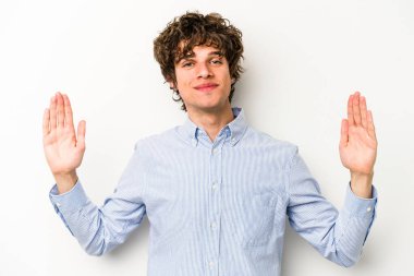 Beyaz arka planda izole edilmiş genç beyaz bir adam işaret parmaklarıyla küçük bir şey tutuyor, gülümsüyor ve kendinden emin..