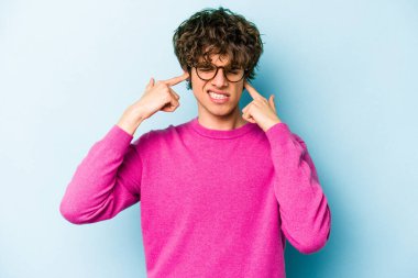 Genç beyaz adam mavi arka planda izole edilmiş parmaklarıyla kulaklarını kapatıyor, gürültülü bir ortam tarafından stresli ve umutsuz..