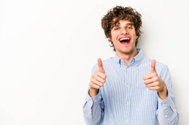 Beyaz arka planda izole edilmiş genç beyaz adam gülümsüyor ve başparmağını kaldırıyor.