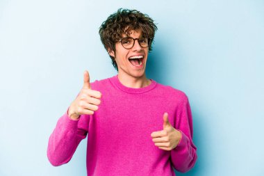 Kafkasyalı genç adam mavi arka planda izole edilmiş. Baş parmağını kaldırıyor, gülümsüyor ve kendinden emin..