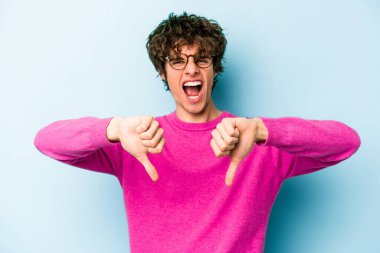 Mavi arka planda izole edilmiş genç beyaz bir adam baş parmağını aşağı gösteriyor ve hoşlanmadığını ifade ediyor..