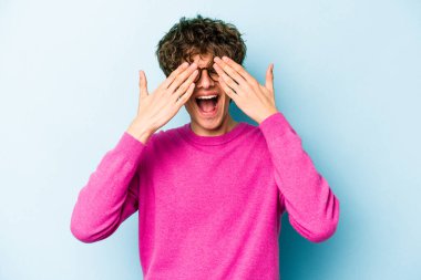 Mavi arka planda soyutlanmış genç beyaz bir adam elleriyle gözlerini kapatıyor, geniş bir gülümsemeyle bir sürpriz bekliyor..