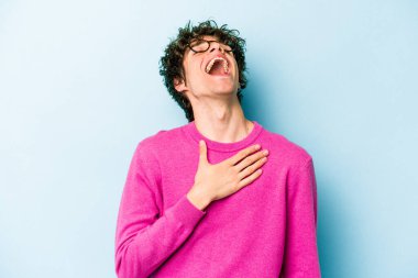 Mavi arka planda izole olmuş genç beyaz adam yüksek sesle gülüyor ve elini göğsüne koyuyor..