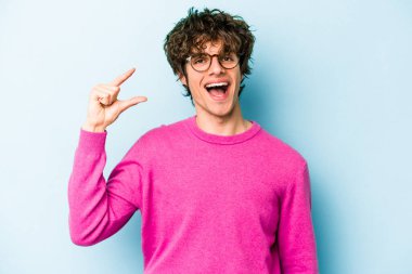 Kafkasyalı genç adam mavi arka planda izole edilmiş. Elinde işaret parmaklarıyla küçük bir şey tutuyor, gülümsüyor ve kendinden emin..