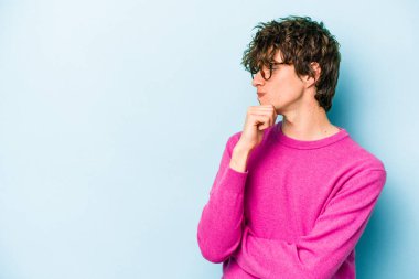 Mavi arka planda izole edilmiş genç beyaz adam şüpheli ve şüpheci bir ifadeyle yana bakıyor..