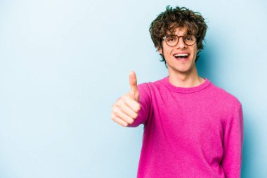 Mavi arka planda izole edilmiş genç beyaz adam gülümsüyor ve başparmağını kaldırıyor.