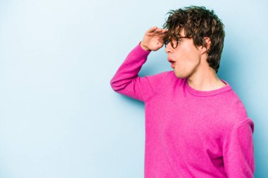 Kafkasyalı genç adam mavi arka planda izole edilmiş, uzaklara bakıyor ve elini alnına koyuyor..