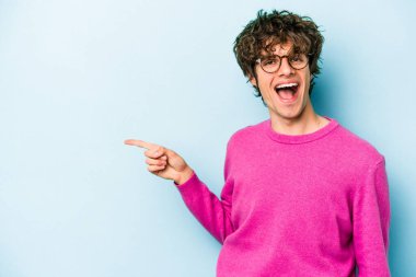 Mavi arka planda izole edilmiş genç beyaz adam işaret parmağıyla işaret ederek gülümsüyor..