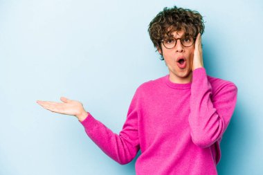 Mavi arka planda izole edilmiş genç beyaz adam, avuç içi fotokopi alanını elinde tutmaktan etkilenmiş..