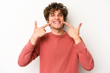 Beyaz arka planda izole edilmiş genç beyaz adam gülümsüyor, parmaklarını ağıza doğrultuyor..
