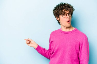 Mavi arka planda izole edilmiş genç beyaz adam gülümsüyor ve boş alanda bir şeyler gösteriyor..