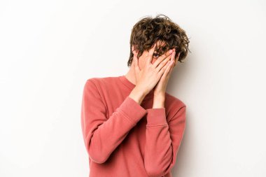 Beyaz arka planda izole edilmiş genç beyaz bir adam kameraya parmaklarıyla göz kırpıyor, yüzünü kapatmaktan utanıyor..