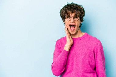 Mavi arka planda izole edilmiş genç beyaz adam yüksek sesle bağırıyor, gözleri açık ve elleri gergin tutuyor..
