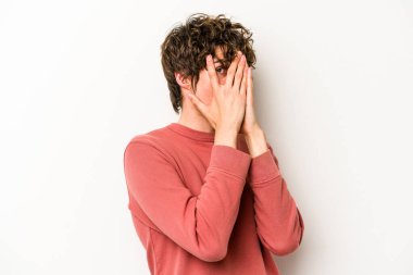 Beyaz arka planda izole edilmiş genç beyaz bir adam parmaklarının arasından korkmuş ve gergin bir şekilde göz kırpıyor..