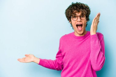 Mavi arka planda izole edilmiş genç beyaz bir adam avucunun içinde fotokopi alanı tutuyor, el üstünde tutuyor. Şaşırmış ve mutlu..